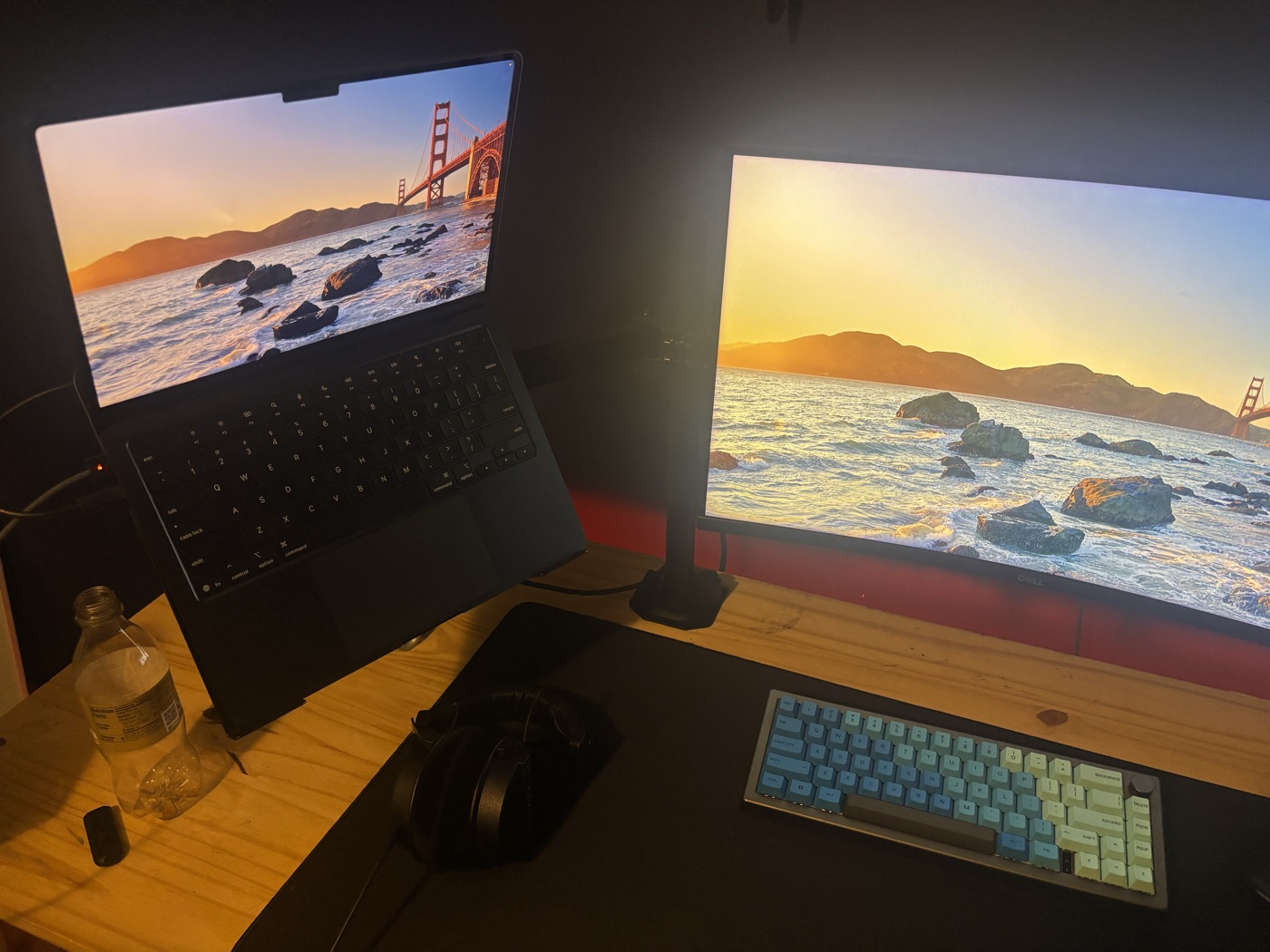 Sam's desk: a MacBook on a stand, a curved monitor, a mechanical keyboard, and headphones on a pine desk with a red backlight